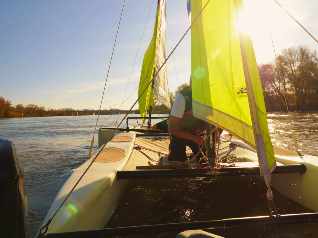 un homme dans un catamaran qui ajuste les cordes de la voile, un rayon de soleil traverse la voile