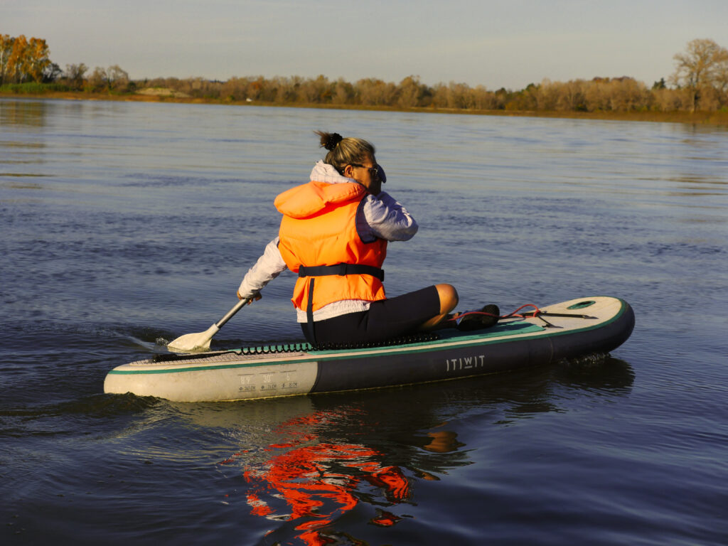 Femme qui fait du paddle sur le Rhône en position assise, elle porte un gilet de sauvetage orange.